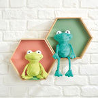 Two plush frogs in hexagonal frames against a white brick wall.