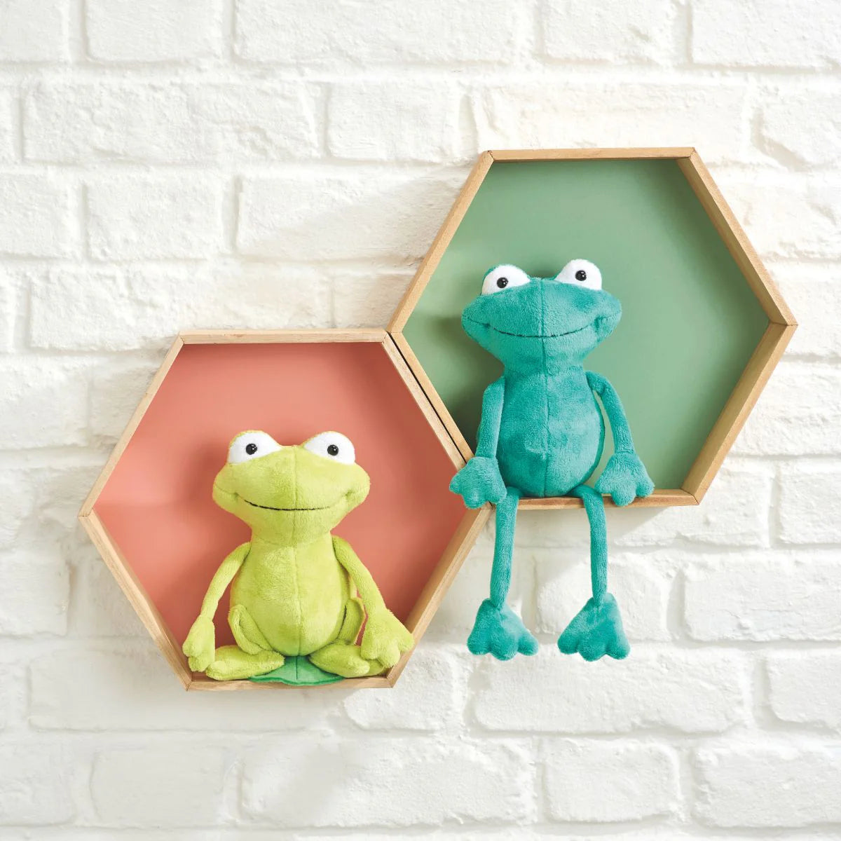 Two plush frogs in hexagonal frames against a white brick wall.