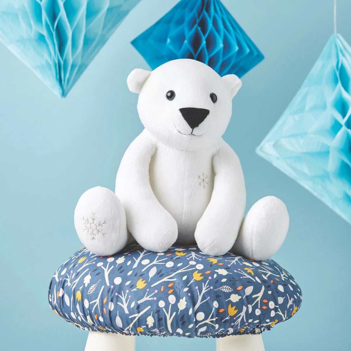White teddy bear sitting on a floral-patterned stool against a blue background with decorative elements.