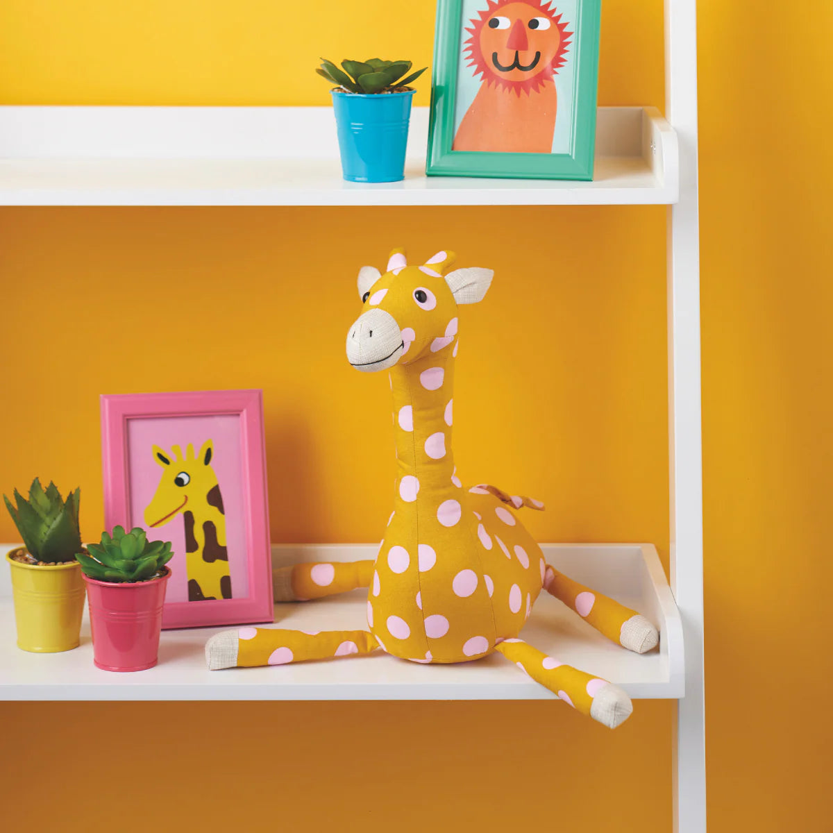 Yellow plush giraffe toy on a white shelf against a yellow wall with framed pictures and plants.