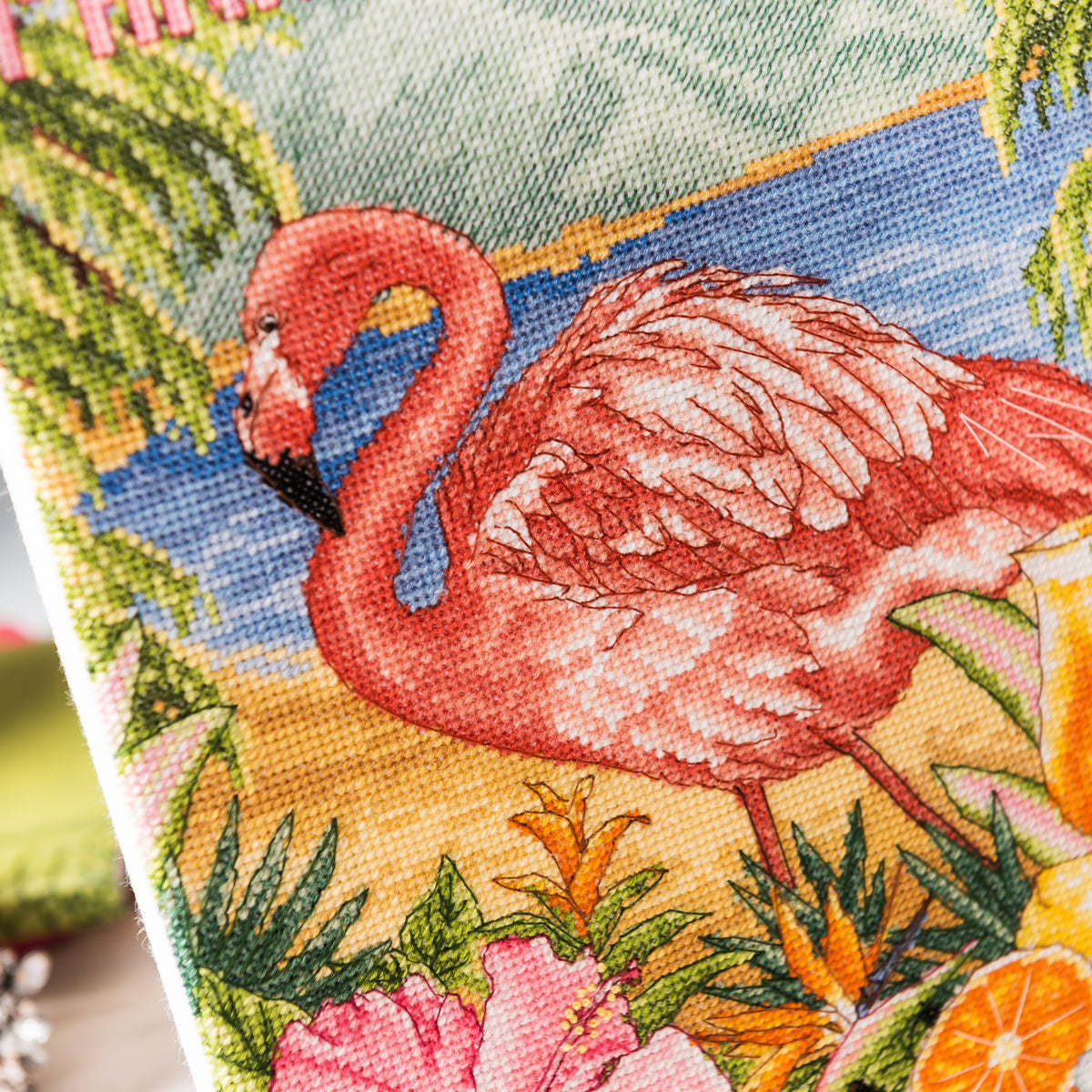 Cross-stitched flamingo design with tropical plants and flowers on fabric.