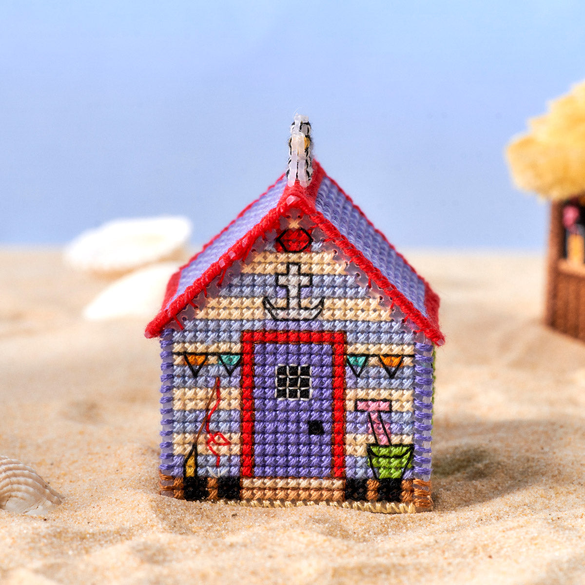Cross-stitched beach hut on a sandy surface with a clear blue sky.