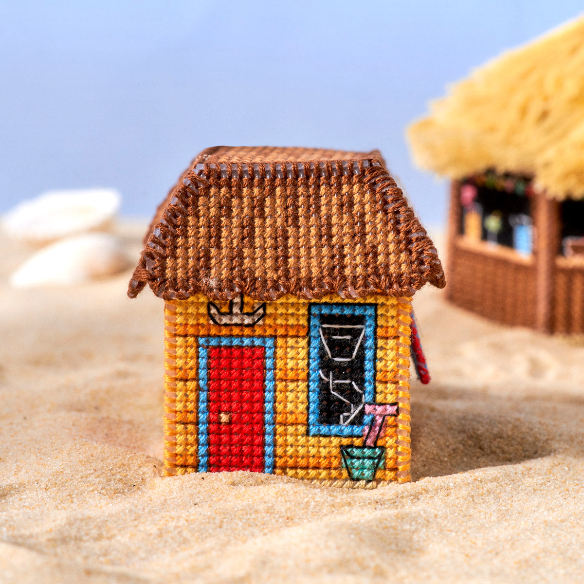 Miniature cross-stitched beach hut on sand with a clear blue sky