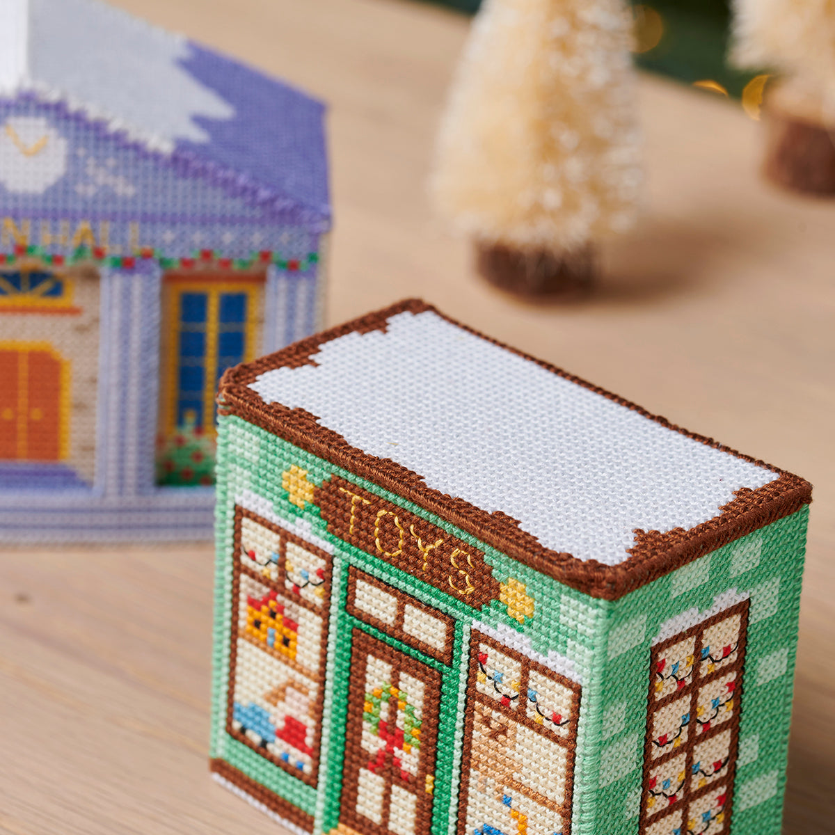 Cross-stitched model of a small building with snow on the roof, placed on a wooden surface.