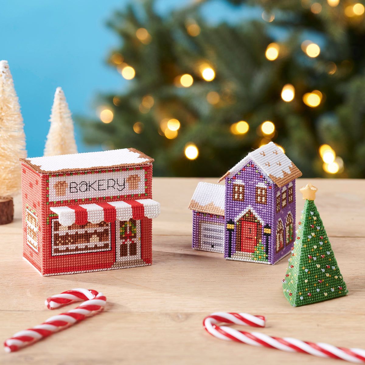Decorative Christmas village scene with a bakery and houses on a wooden surface, blurred Christmas tree in the background.