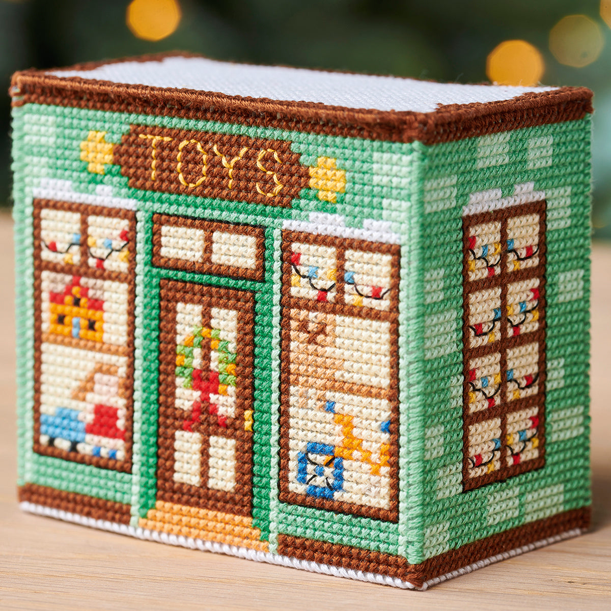 Cross-stitched toy store box with a green facade and brown trim on a wooden surface.
