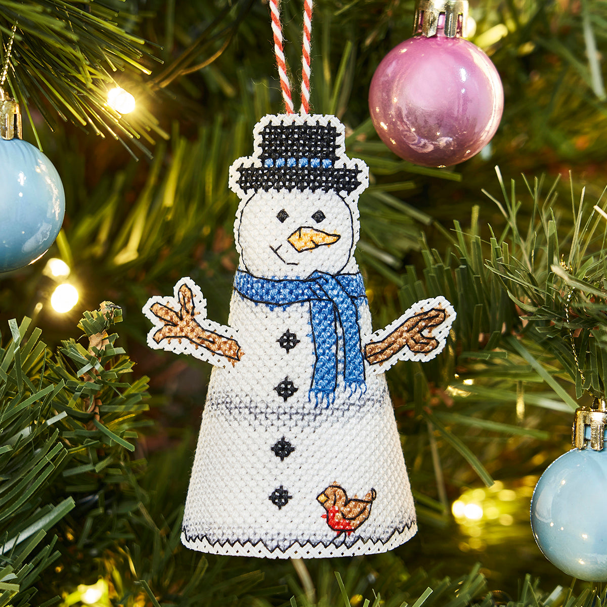 Cross-stitched snowman ornament on a Christmas tree with colorful ornaments.