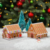 Two cross stitch gingerbread house ornaments on a snowy surface with Christmas trees and decorations in the background.