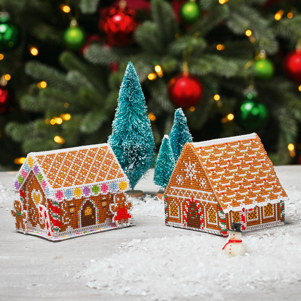 Two cross stitch gingerbread house ornaments on a snowy surface with Christmas trees and decorations in the background.