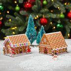 Two cross stitch gingerbread house ornaments on a snowy surface with Christmas trees and decorations in the background.
