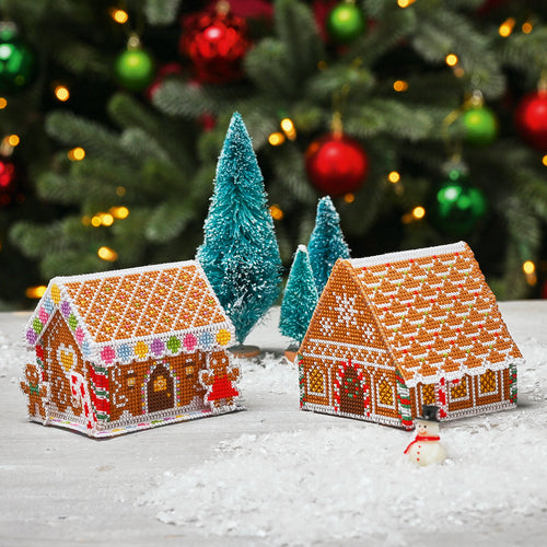 Two cross stitch gingerbread house ornaments on a snowy surface with Christmas trees and decorations in the background.