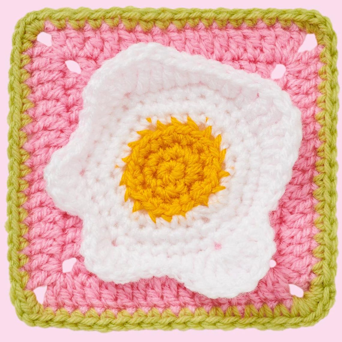 Crochet square featuring a sunny-side-up egg design. The yolk is yellow, surrounded by white, set on a pink background with a green border. Playful and cozy.