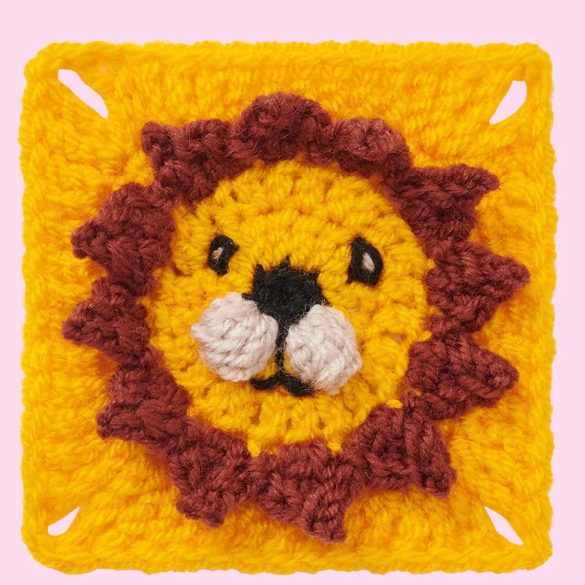 A crocheted square with a cute lion face in the center. The lion has a round yellow face, brown mane, black eyes, and a pink background. Playful and cheerful.