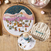 Embroidery hoop with mountain scene, knitted item, and mug with embroidered design on wooden surface