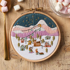 Embroidery hoop with a winter landscape design on a wooden surface with marshmallows and a spoon.