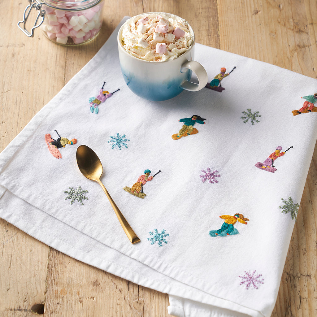 White napkin with embroidered figures and snowflakes on a wooden surface with a mug of hot chocolate.