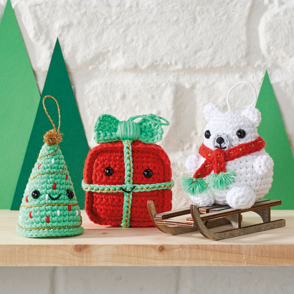 Crocheted Christmas ornaments on a shelf: a smiling tree, a red gift with a bow, and a polar bear wearing a red scarf on a wooden sled. Festive and cheerful.