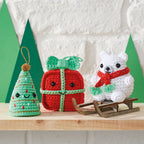 Crocheted Christmas ornaments on a shelf: a smiling tree, a red gift with a bow, and a polar bear wearing a red scarf on a wooden sled. Festive and cheerful.