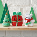 A crochet pattern set featuring a Christmas tree, a gift box, and a polar bear amigurumi. The Christmas tree is green with a gold ornament, the gift box is red with a green ribbon, and the polar bear is white with a red scarf, all three are adorned with pom-poms.