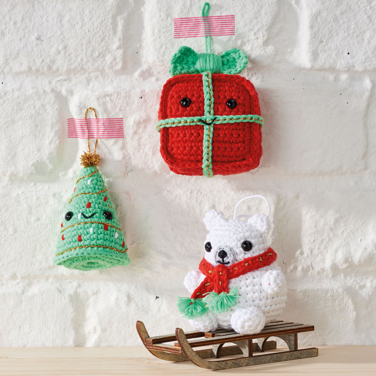 Crocheted Christmas ornaments on a white wall, including a red gift with green bow, a smiling green tree, and a white bear on a wooden sled.