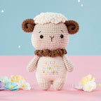 Cute crocheted lamb with brown ears and a fluffy white top, standing on a pink surface. Scattered colorful flowers and a pastel blue background with stars add whimsy.