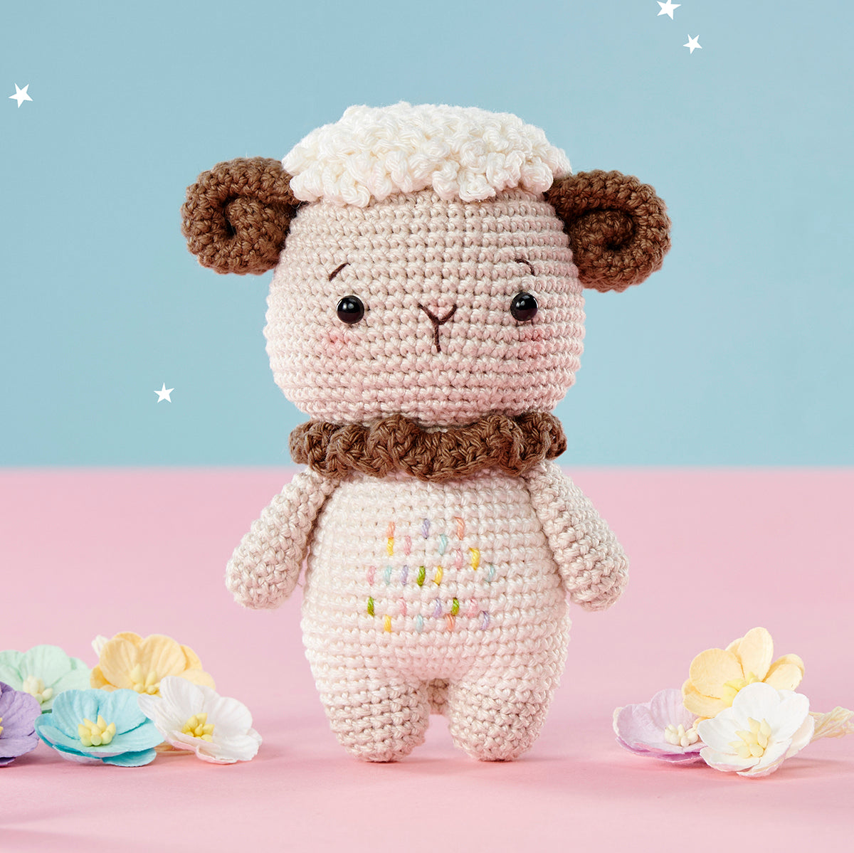 Cute crocheted lamb with brown ears and a fluffy white top, standing on a pink surface. Scattered colorful flowers and a pastel blue background with stars add whimsy.