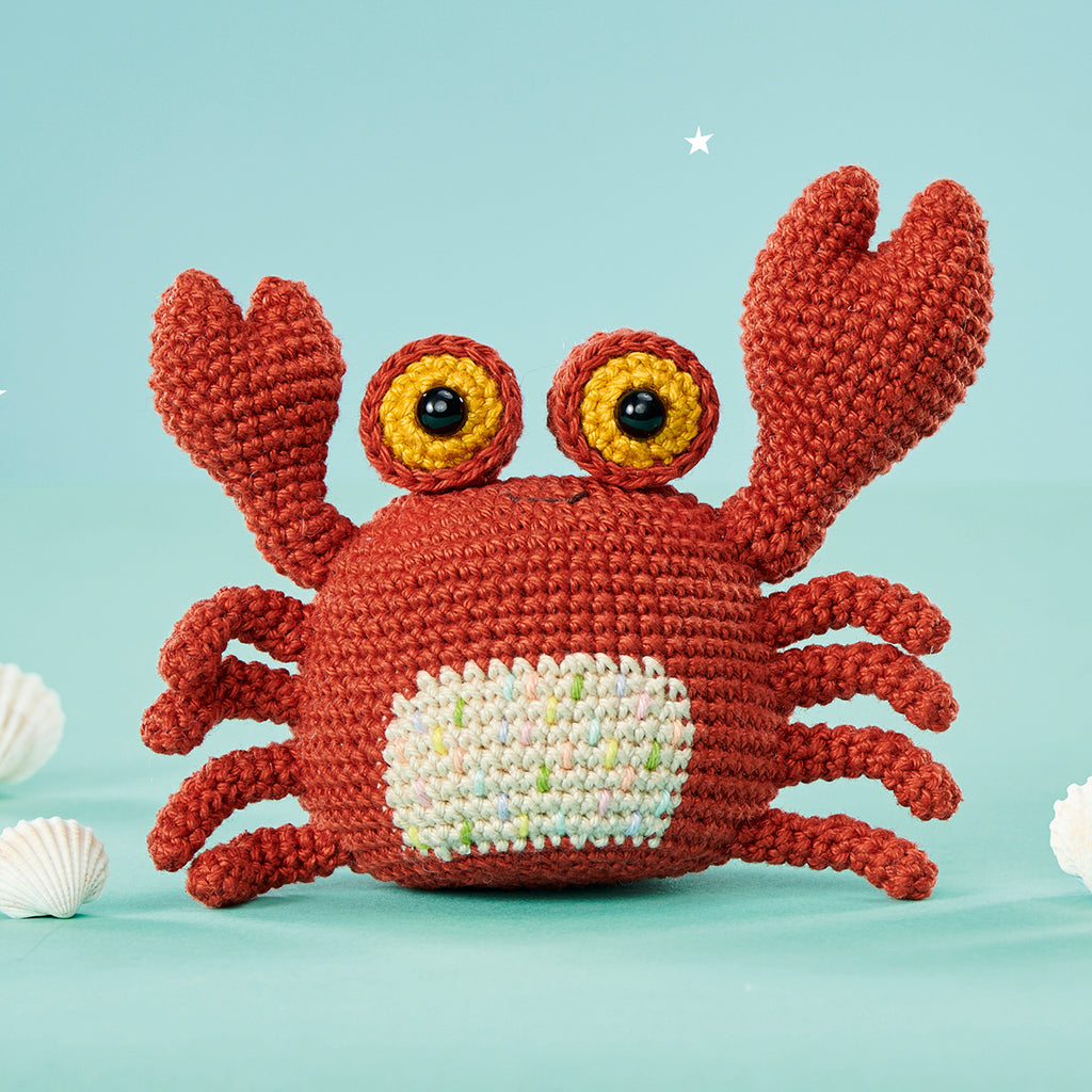 Crocheted red crab with expressive yellow eyes on a pastel blue background. Two white seashells are placed beside it, creating a cheerful, whimsical feel.