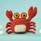 Crocheted red crab with expressive yellow eyes on a pastel blue background. Two white seashells are placed beside it, creating a cheerful, whimsical feel.