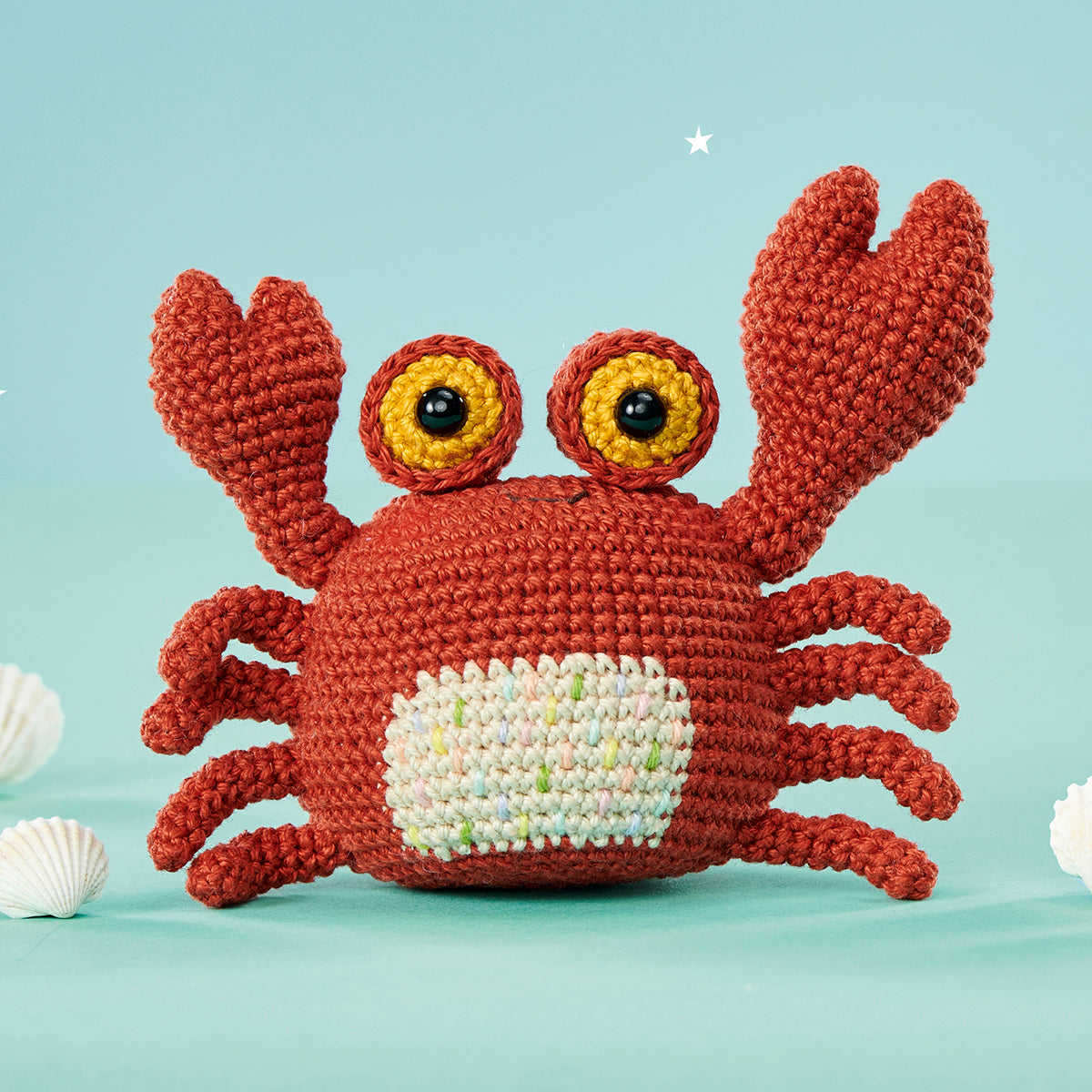 Crocheted red crab with expressive yellow eyes on a pastel blue background. Two white seashells are placed beside it, creating a cheerful, whimsical feel.