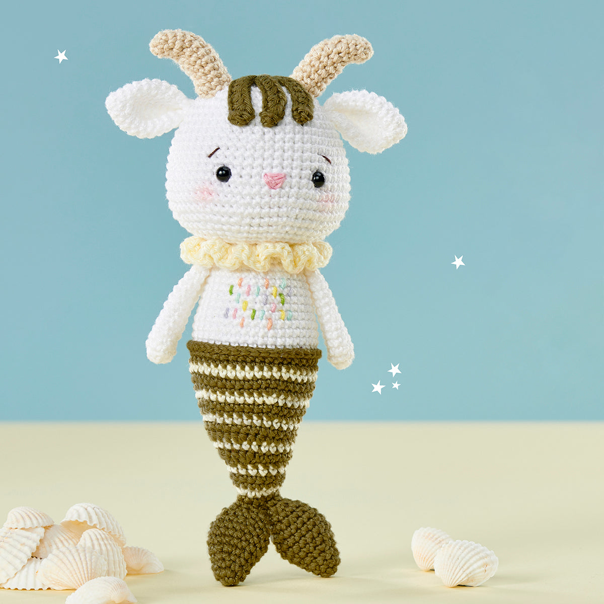 Crocheted goat-mermaid hybrid with a striped tail, cute face, and blushing cheeks. Set against a blue backdrop with small stars and seashells nearby.