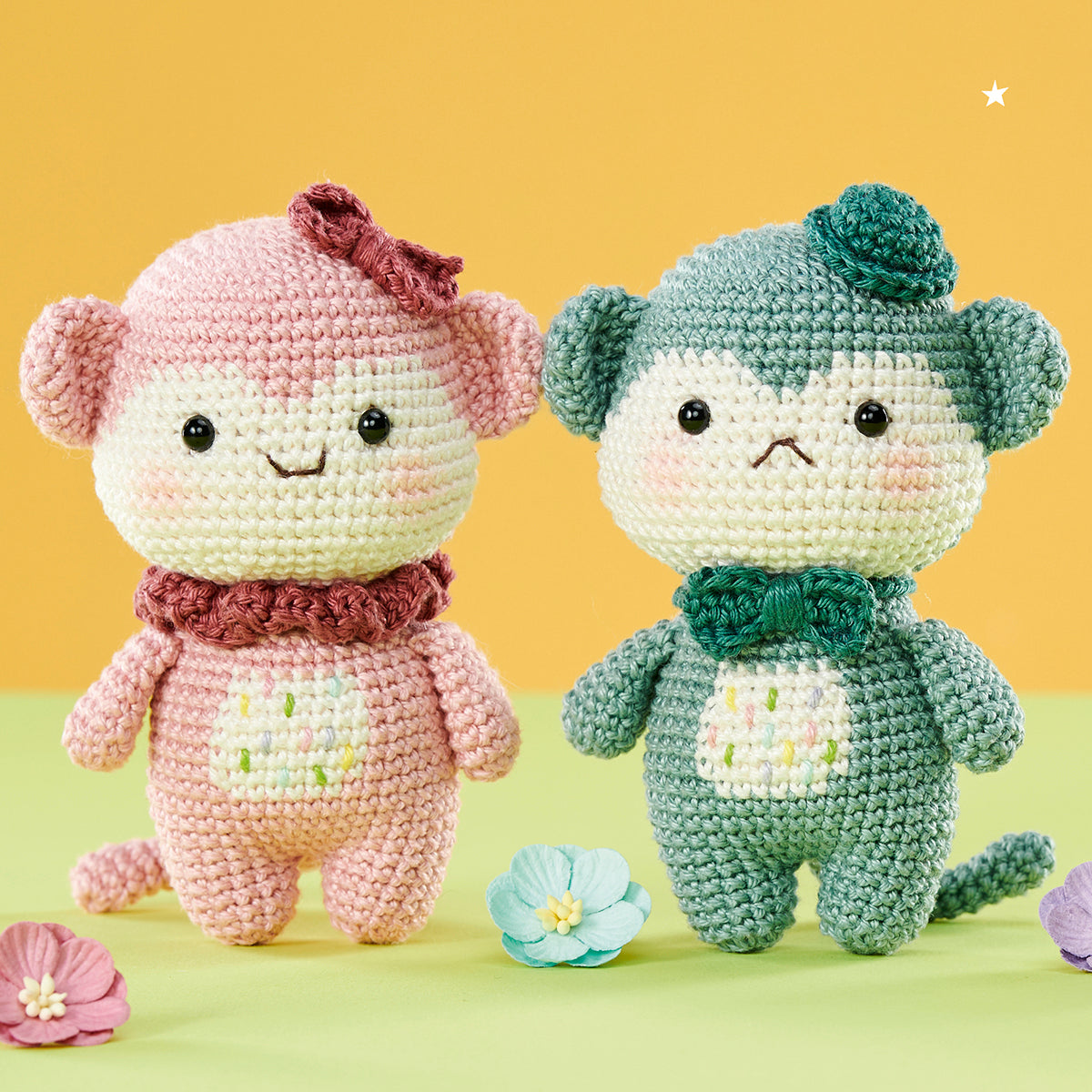 Two crocheted dolls, a pink monkey with a bow and a green monkey with a hat, stand on a pastel surface. Colorful flowers accent the playful scene.