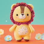 A cute crocheted lion toy with a pink mane stands on an orange surface. It has a brown stripe, small ears, and a stitched belly, with pastel flowers nearby.