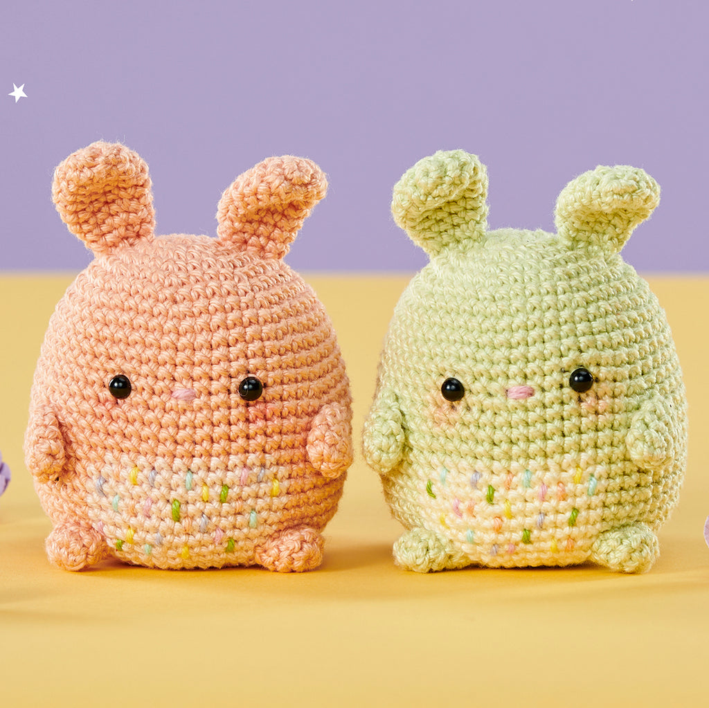 Two amigurumi bunnies, one peach and one green, with round black eyes, sit side by side on a yellow and purple background, evoking a cute, cheerful vibe.