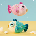 Two crocheted fish toys, one pink with purple accents and one green with blue accents, float on a pastel background with small seashells nearby. Playful and whimsical.