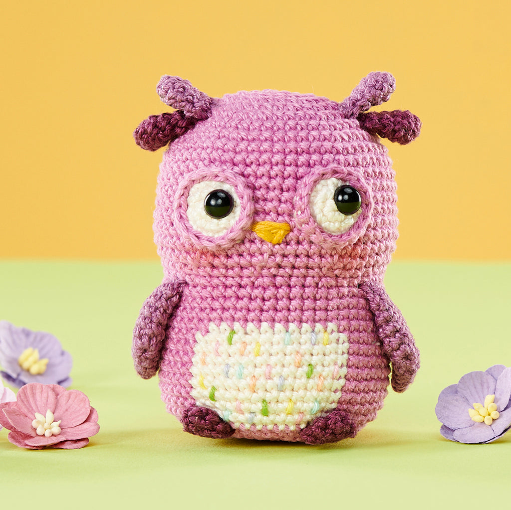 A cute, pink crocheted owl with large eyes and a yellow beak sits surrounded by pastel flowers on a green and orange background, evoking a playful tone.