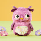 A cute, pink crocheted owl with large eyes and a yellow beak sits surrounded by pastel flowers on a green and orange background, evoking a playful tone.