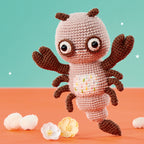 Cute crocheted scorpion with large eyes and brown claws against a teal background. It's surrounded by small white and yellow flowers on an orange surface.