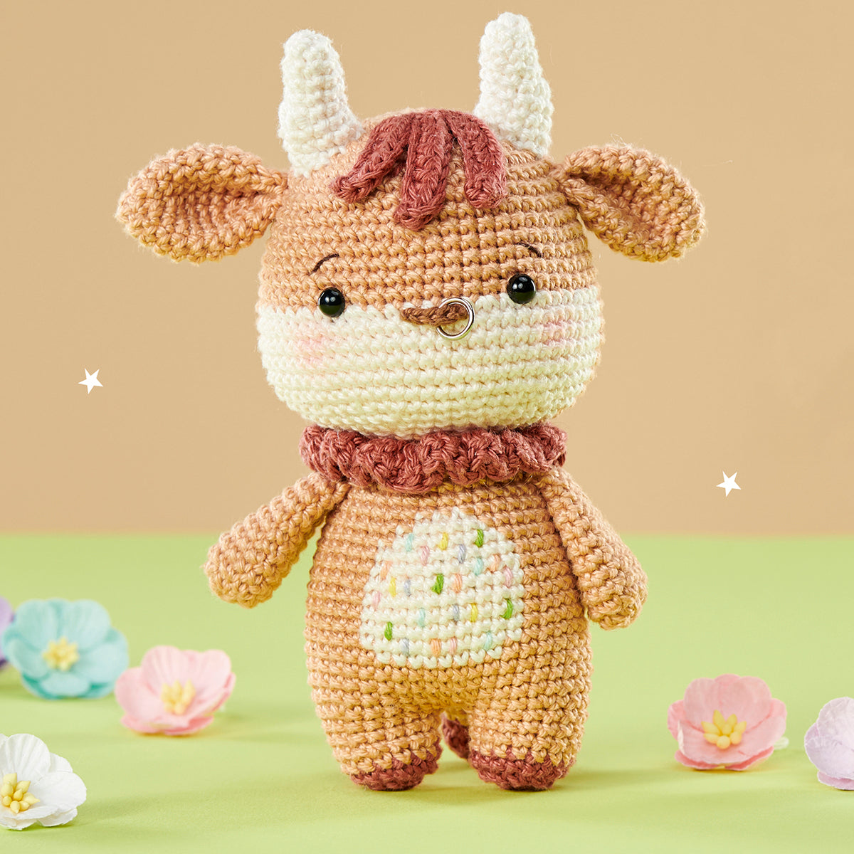 Cute crocheted bull doll with beige body, pink nose, and a ring in its snout stands on green surface. Colorful pastel flowers surround it.