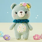 A crochet pattern image featuring a Stuffed toy bear with a blue scarf and floral patterns, surrounded by colorful flowers.