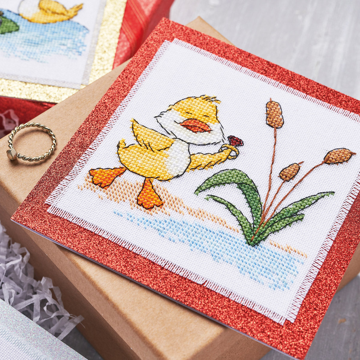 Cross-stitched duck design on a red border with a gift box in the background