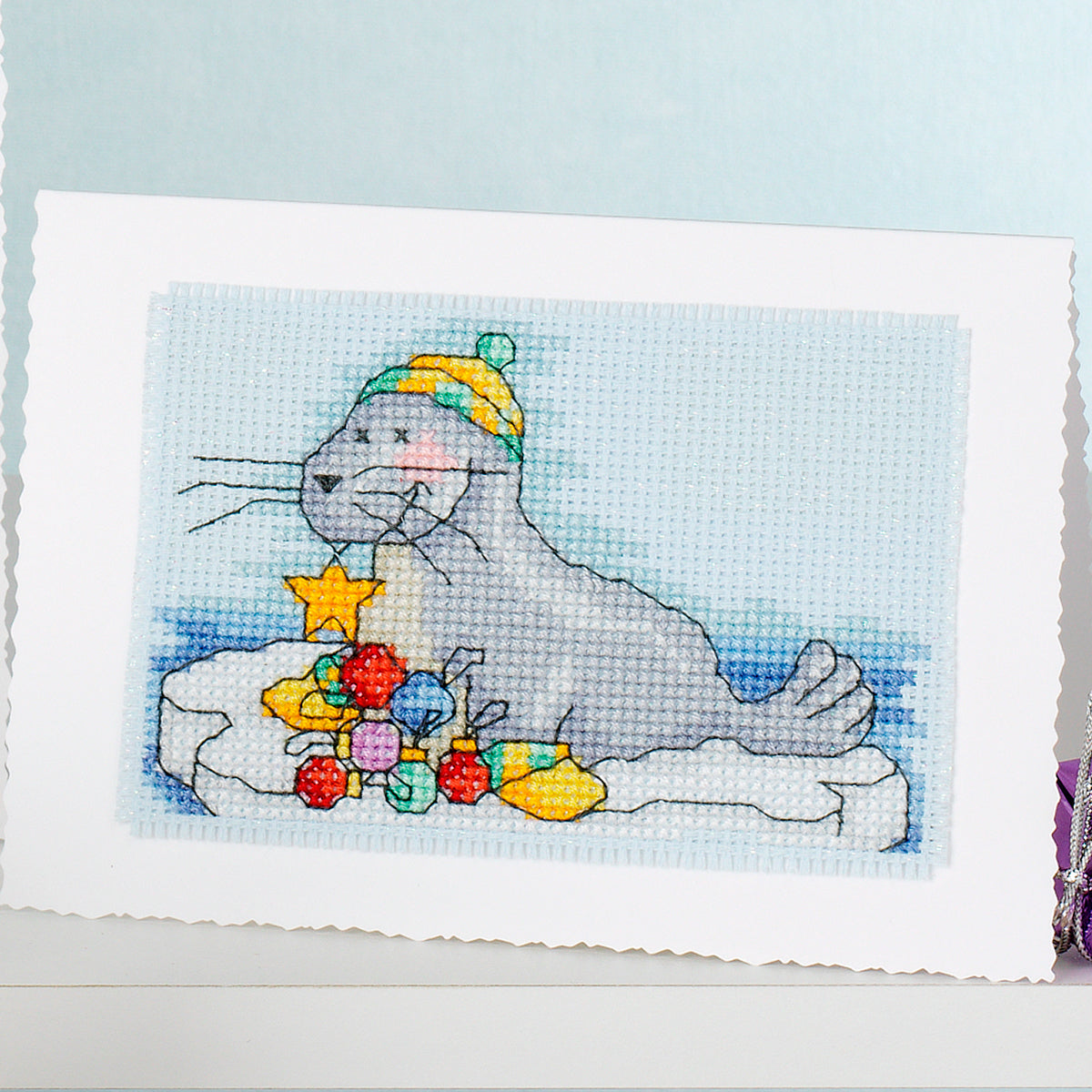 Cross-stitched design of a seal with colorful toys on a light blue background
