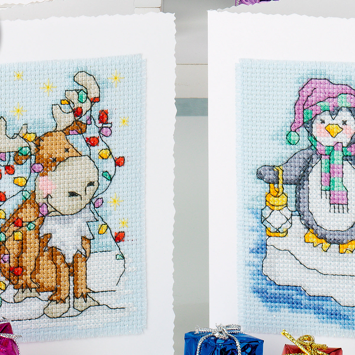Two cross-stitched pictures of a deer and a penguin with Christmas decorations on a white background.