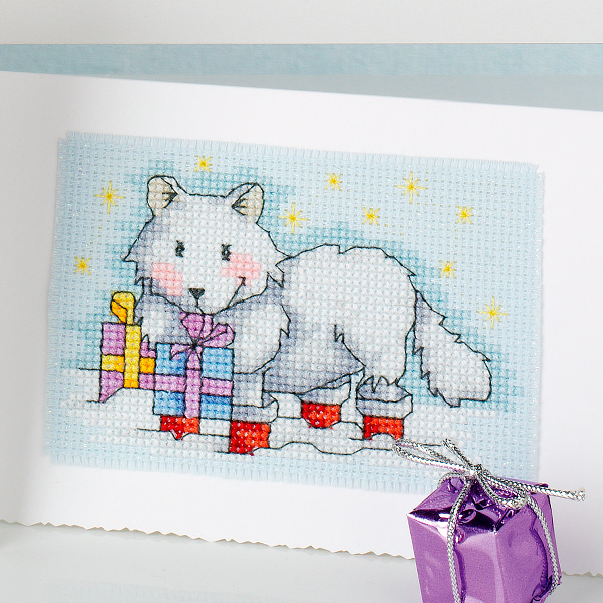 Cross-stitched design of a white fox with presents on a blue background, accompanied by a purple gift box.