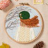Embroidery hoop with a person wearing a green hat and plaid scarf against a blue background.
