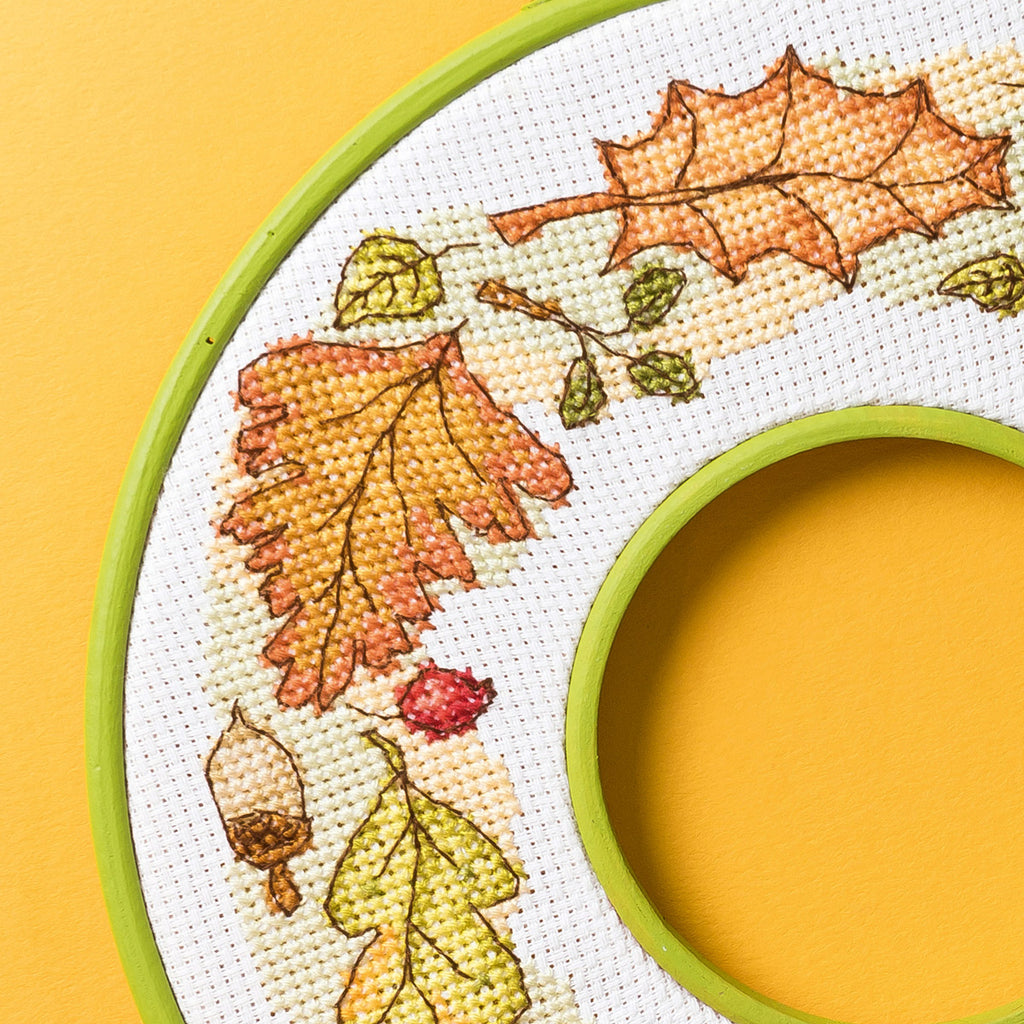 Cross-stitched design of autumn leaves and acorns in an embroidery hoop on a yellow background.