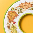 Cross-stitched design of autumn leaves and acorns in an embroidery hoop on a yellow background.