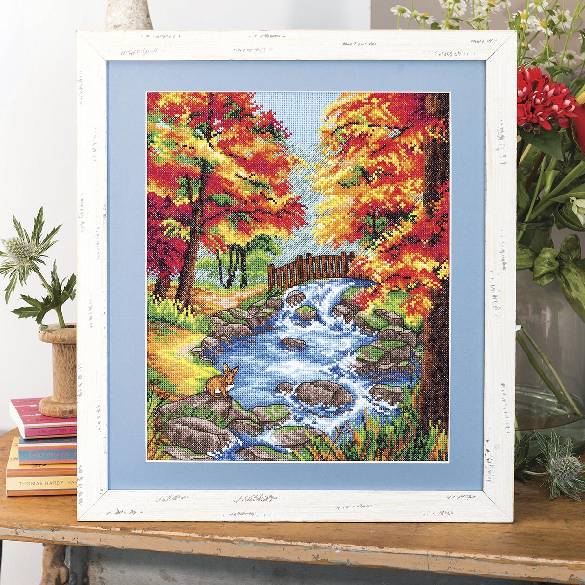Framed cross-stitched picture of a autumn landscape with trees, stream, and deer.