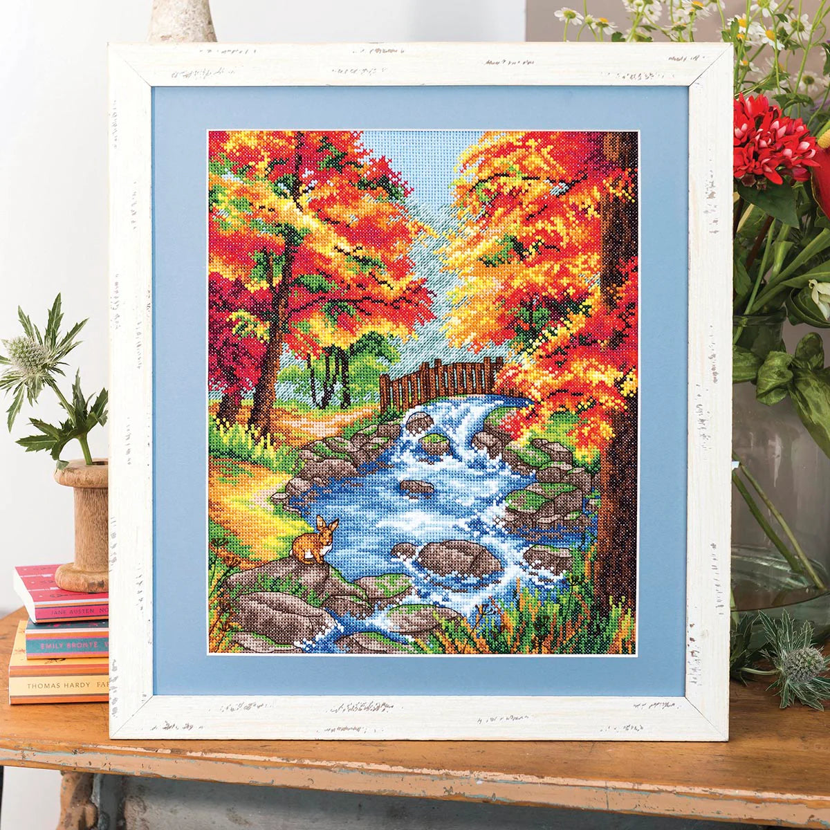 Framed cross-stitch artwork of a autumn landscape with trees, a stream, and a fox.