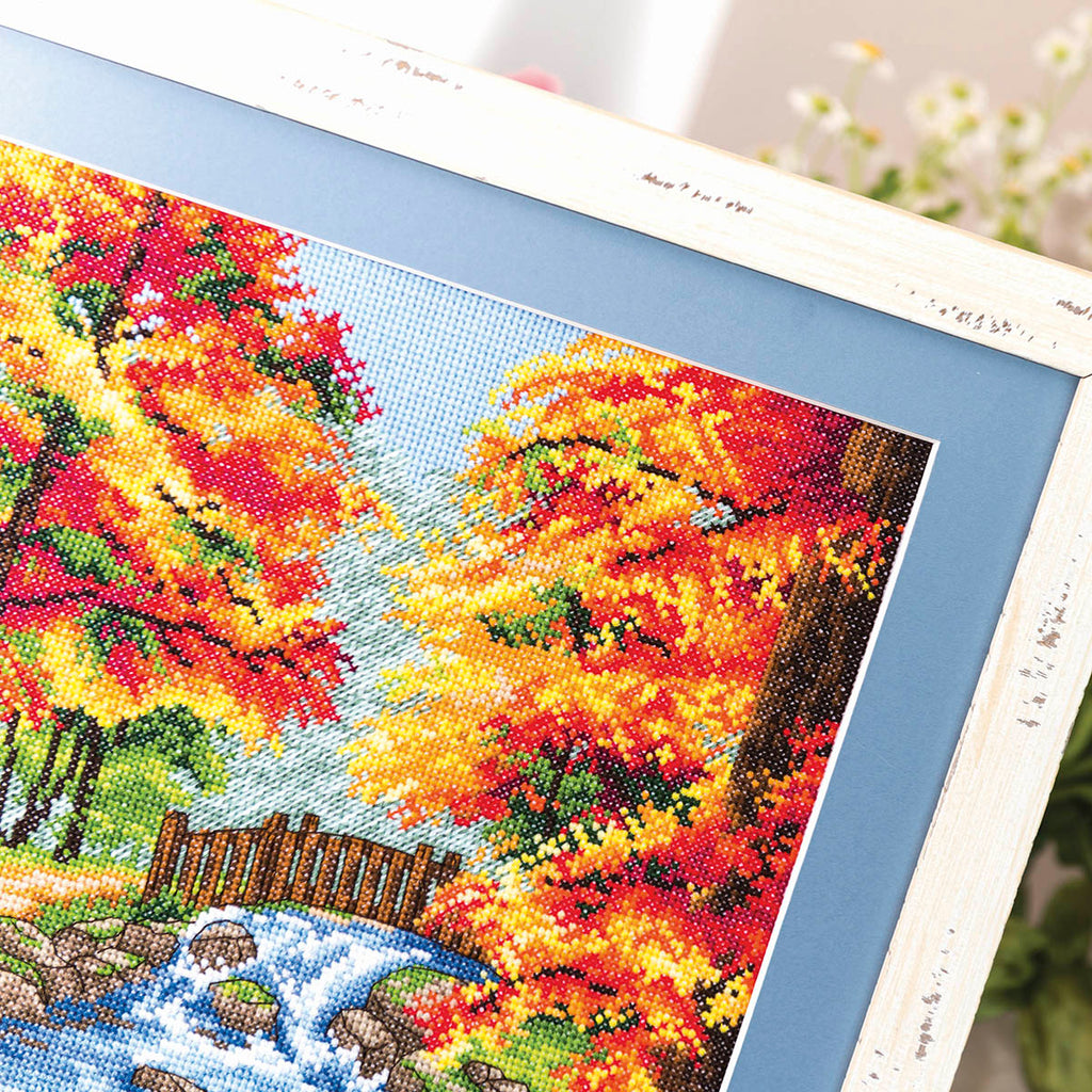 Cross-stitched scene of autumn trees with a stream and wooden fence.