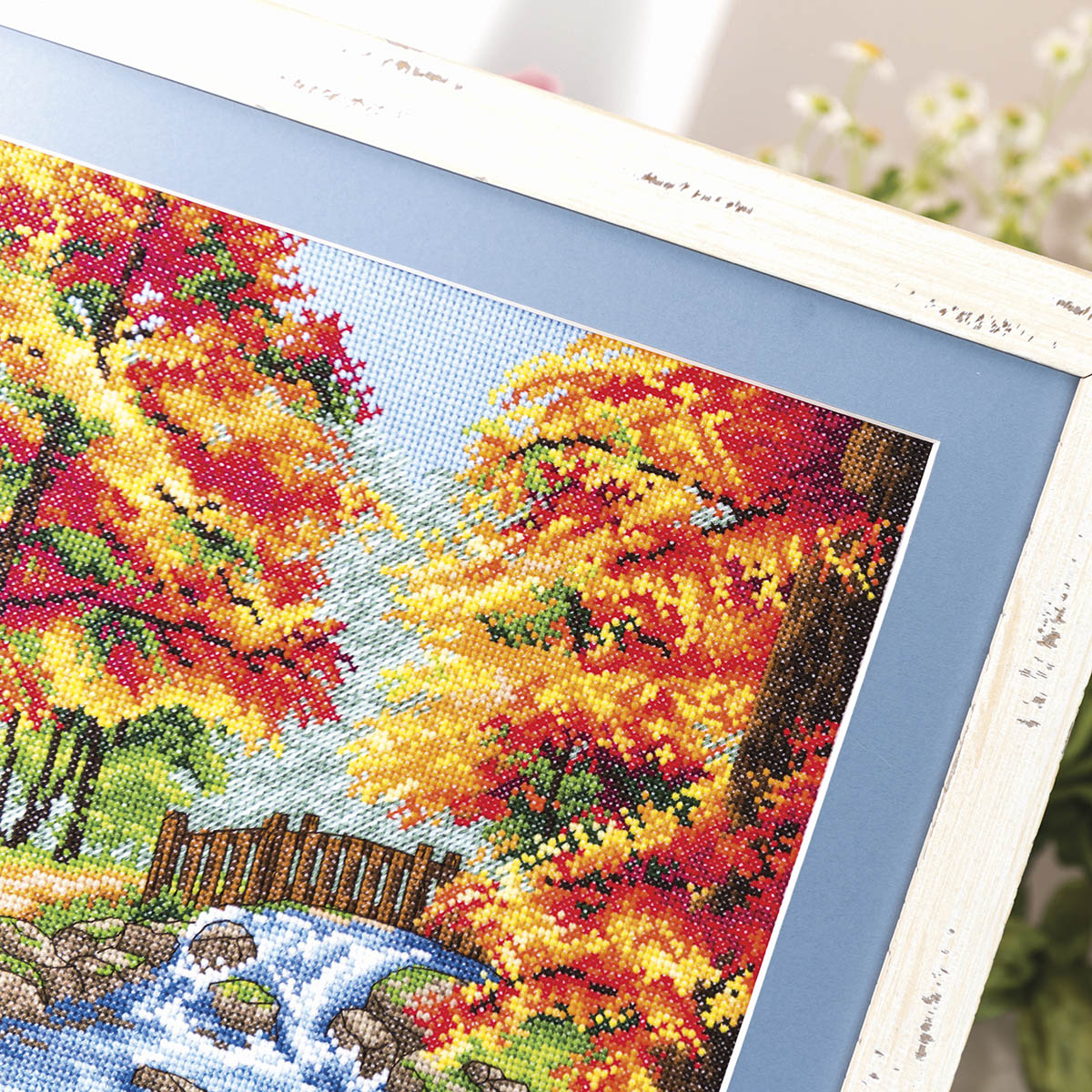 Cross-stitched scene of autumn trees with a stream and wooden fence.